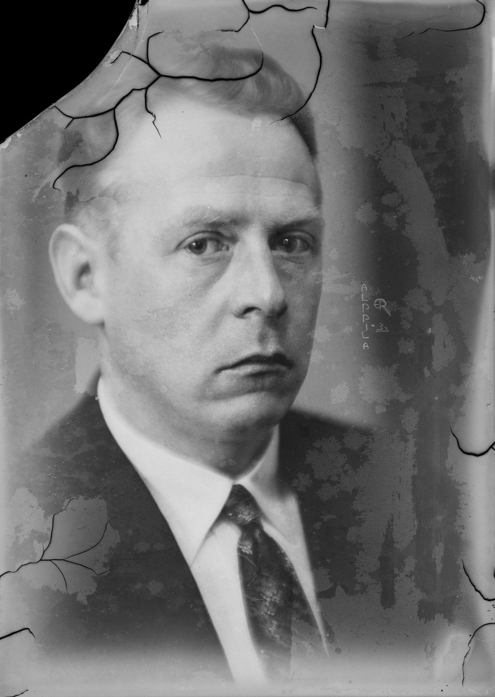 A black-and-white studio portrait of a man, cracked and stained, with the top corner of the print missing